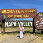 Couple kissing in front of the iconic “Welcome to this world famous wine growing region – Napa Valley” sign on a sunny day, with vineyards and mountains in the background.