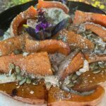 Roasted honey nut squash wedges arranged on a platter over a smooth white bean purée, topped with a toasted pumpkin seed and spice crunch, garnished with flowering purple basil