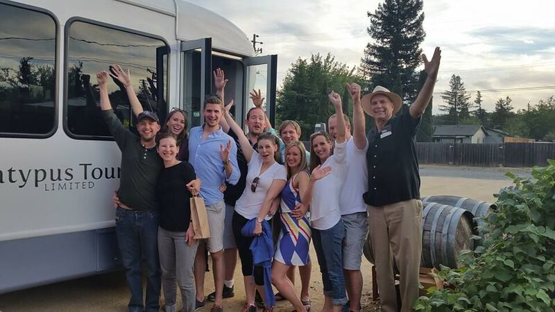 A group of people poses joyfully outside a tour bus, waving and smiling, with trees and barrels in the background.