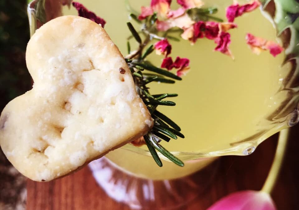 A close-up of a delicate yellow-green shrub cocktail garnished with dried rose petals and a sprig of rosemary, served in a scalloped glass. A heart-shaped shortbread cookie is perched on the rim, and a pink tulip rests nearby on the wooden surface, adding a romantic touch.