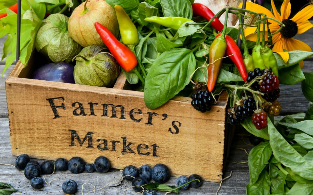 Wooden crate labeled "Farmer's Market" filled with fresh produce, including tomatillos, chili peppers, basil, blackberries, and garlic, with blueberries scattered on a rustic wooden surface.