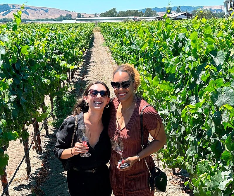 Teo people on the middle of a vinetyard row with a building and hills in the background
