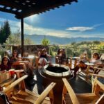 A small group of women relaxing around a firepit on a sunny winery deck, raising their glasses toward the camera with vineyard and mountain views behind them.