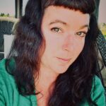 Portrait of Courtney, a Platypus Tours guide, smiling while seated on a tour bus, with long dark hair and a green top, vineyard scenery visible through the window behind her.
