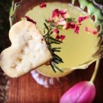 A close-up of a delicate yellow-green shrub cocktail garnished with dried rose petals and a sprig of rosemary, served in a scalloped glass. A heart-shaped shortbread cookie is perched on the rim, and a pink tulip rests nearby on the wooden surface, adding a romantic touch.