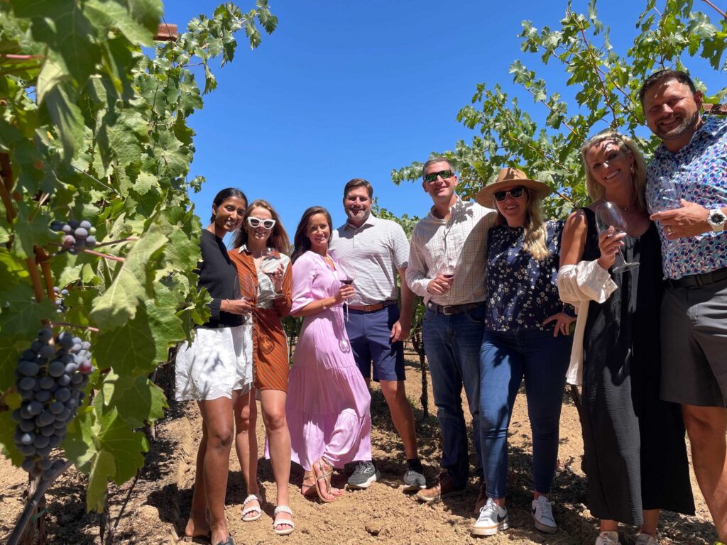 Guests on a wine tour standing among vineyard rows, smiling and holding wine glasses under a clear blue sky.