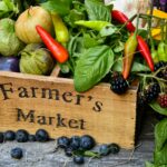 Wooden crate labeled "Farmer's Market" filled with fresh produce, including tomatillos, chili peppers, basil, blackberries, and garlic, with blueberries scattered on a rustic wooden surface.