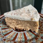 Slice of pumpkin spice cheesecake topped with whipped cream and sprinkled with nutmeg, served on a decorative patterned plate.