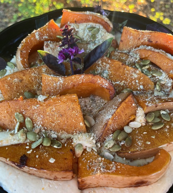 Roasted honey nut squash wedges arranged on a platter over a smooth white bean purée, topped with a toasted pumpkin seed and spice crunch, garnished with flowering purple basil