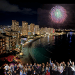 Group of people wearing leis celebrating along Waikiki Beach at night with city lights and fireworks over Honolulu, representing Platypus Perks travel experiences in Hawaii.