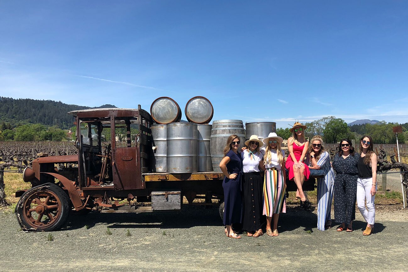 Wine tour guests posing beside a vintage truck loaded with wine barrels in a vineyard setting.