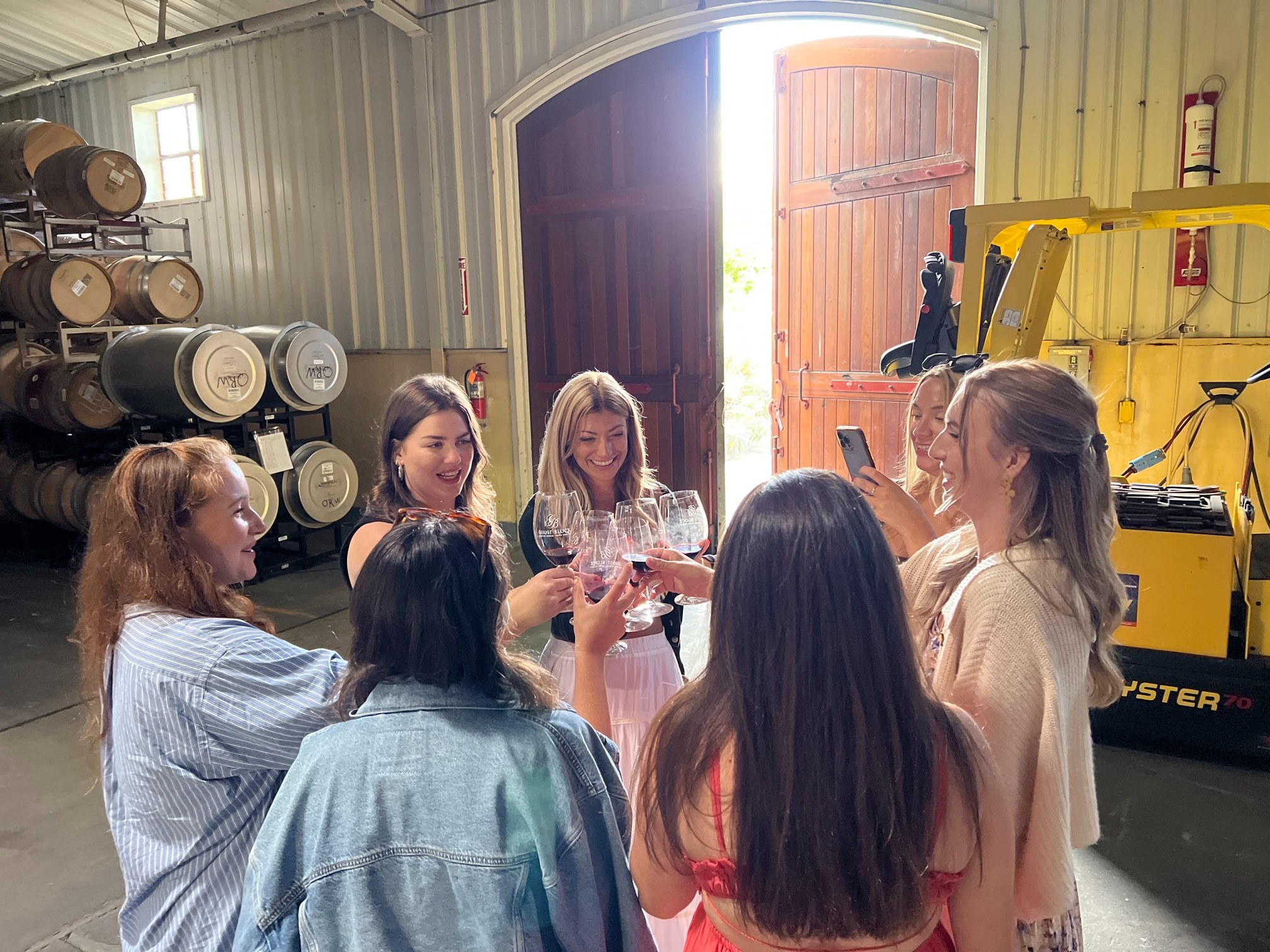 Six women raising their wine glasses in a toast inside a winery production room, surrounded by stacked oak barrels.