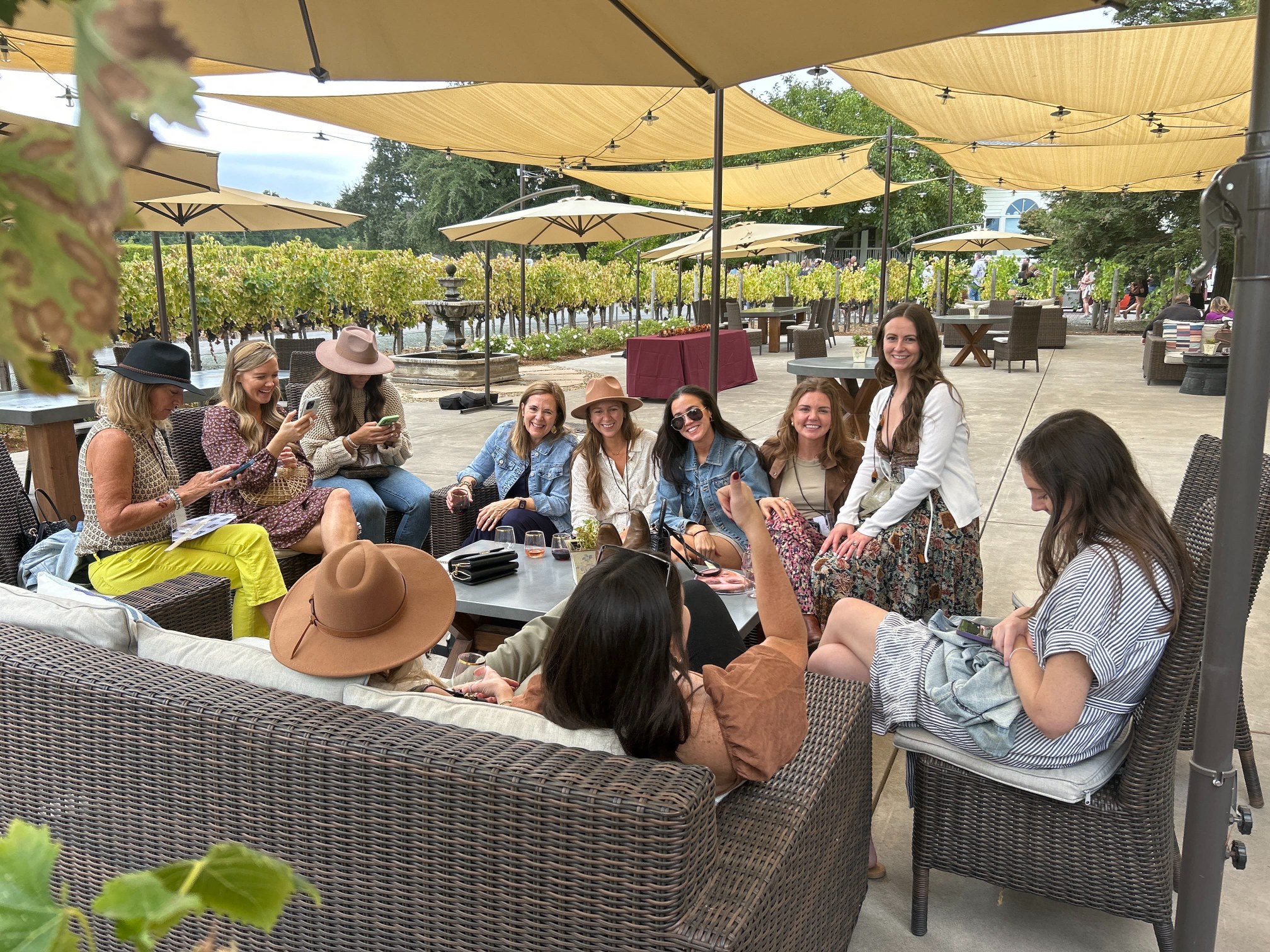 Family and friends enjoying a wine tasting together on a shaded winery patio, surrounded by grapevines and outdoor seating.