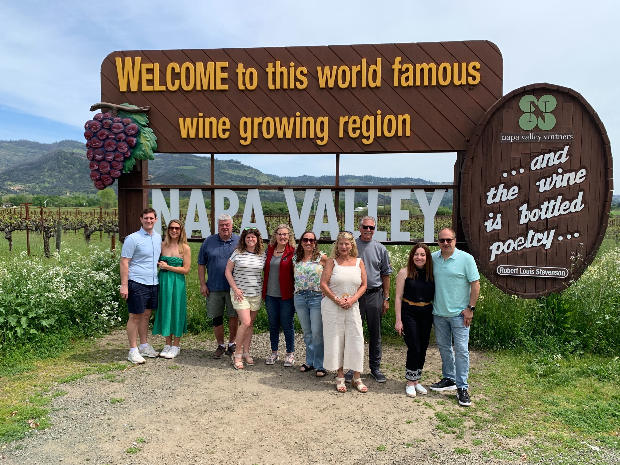 Platypus Wine Tours guests posing together in front of the iconic Welcome to Napa Valley sign, with vineyards and mountains in the background.