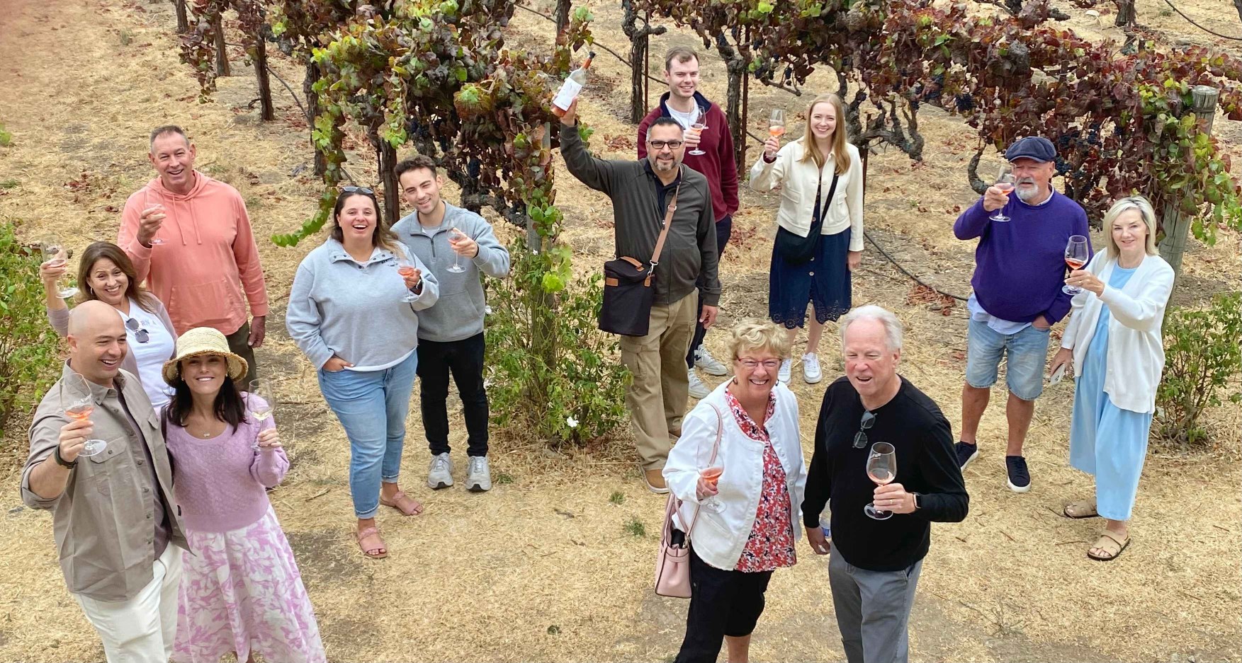 Platypus Wine Tours guests smiling, waving, and raising their wine glasses together among the grapevines during a wine country tour.