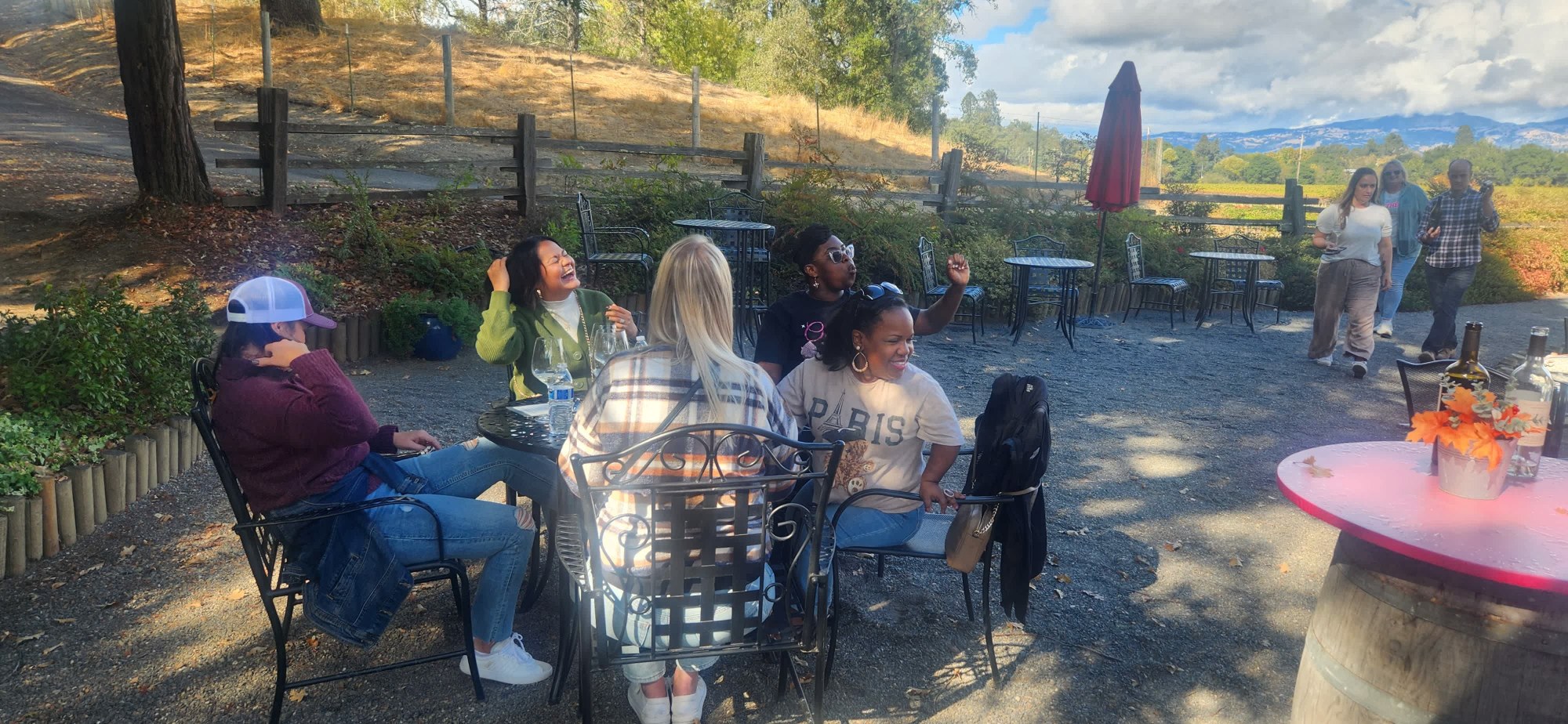Platypus Wine Tours guests relaxing and chatting around an outdoor winery patio table, surrounded by trees and scenic wine country hills.