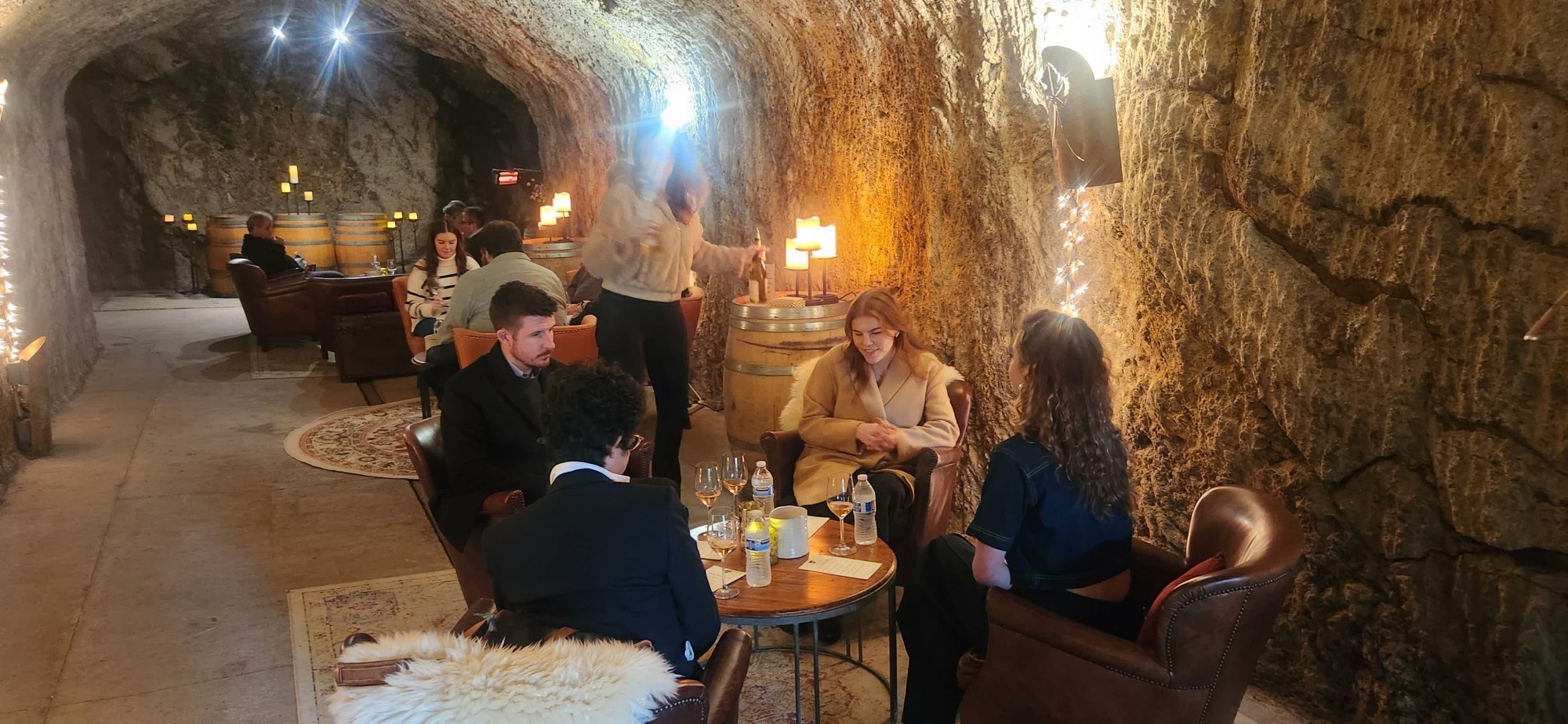 Wine tour guests enjoying an intimate wine tasting in a warmly lit, stone-walled winery cave with comfortable lounge seating.