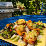 Grilled Hawaiian chicken served over coconut white rice with warm pineapple topping, fresh salad, lime wedges, and edible flowers on a blue patio table beside a backyard grill.